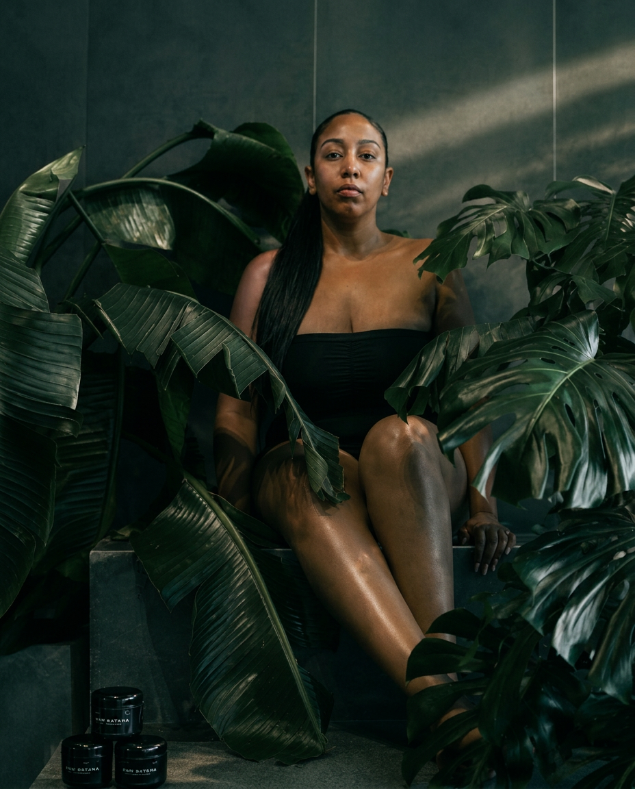 Sarah seated among rainforest leaves with Raw Batana jars